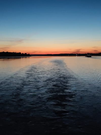 The scene depicts a wide body of water in Il'inskoe, Russia, at twilight. The sky transitions horizontally from deep blue at the zenith, fading to a lighter blue and then a prominent band of orange and peach hues just above the horizon. A dark, silhouetted landmass or treeline extends across the entire horizon, separating the sky from the water. On the far right of this distant landmass, several distinct bright white artificial lights are visible.

The water's surface reflects the sky's colors, showing an orange-peach glow near the horizon, transitioning to dark blue-grey in the mid-ground. A prominent, wide, turbulent wake extends from the mid-ground directly towards the viewer, dominating the lower half of the image. This wake indicates a vessel recently passed or is actively moving away. The water within the wake is heavily textured with ripples and foam, appearing darkest in the immediate foreground, almost black. In the mid-ground, on the right side of the frame, a boat with multiple illuminated white lights is visible, heading towards the right and slightly away from the viewer. Another faint light source is visible further left along the distant horizon. No individuals are discernible. The overall lighting condition is low, indicative of either dawn or dusk.