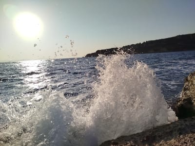 The image captures a dynamic scene of ocean waves crashing against rocky shores. The sun is visible in the upper left quadrant, casting a bright glare on the water, which is a deep blue with ripples reflecting the sunlight. Two large splashes of water are prominently featured in the foreground, frozen in motion as they burst upwards from the impact. The texture of the splashes is crystalline, with countless droplets of varying sizes suspended in the air. In the background, a dark, tree-covered hillside slopes down towards the sea, suggesting a coastal landscape. The sky is a clear, pale blue, with a slight gradient towards the horizon. The rocks in the foreground are rough and textured, appearing to be a natural part of the coastline. The scene is one of natural beauty and powerful ocean activity.