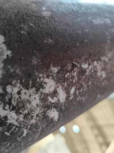 The image displays a close-up, abstract view of a weathered, dark-colored cylindrical surface. The surface exhibits a rough, uneven texture with patches of lighter material, possibly paint or mineral deposits, adhering to it in irregular patterns. Some small, dark specks are visible on the surface, potentially dust or debris. In the lower right portion of the image, a blurred background suggests an outdoor environment with soft, out-of-focus light sources that appear as bright, diffused orbs. The lighting is subdued, creating a slightly hazy or diffused atmosphere. The image is oriented with the surface occupying the majority of the frame, angled diagonally. No people or specific landmarks are identifiable, and the image does not provide information about its location within the city of Краснадворскі сельскі Савет, Belarus.