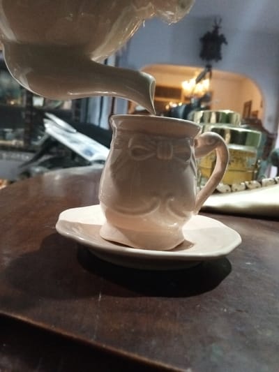 A close-up, low-angle photograph taken indoors in Longmiang, Laos, captures a light-colored ceramic teapot pouring liquid into a matching ceramic cup with saucer. The teapot's spout is visible entering the frame from the upper left, directing a thin stream of liquid downwards into the cup, which is positioned slightly right of center. The cup is pale cream or light pink, featuring an embossed bow design near its rim and scrolled decorative elements below. It has a single handle on its right side and rests on a matching saucer. Both the cup and saucer are situated on a dark brown, possibly wooden, table surface that occupies the lower half of the frame and shows minor surface imperfections. The background is significantly blurred, revealing an indoor environment with artificial lighting. To the right of the cup, indistinct objects with metallic (possibly gold or brass) lids and a light-colored textile are present on the table. Further in the background, a warm-toned light fixture is visible through an arched opening, and a dark, hanging object, possibly a clock, is discernible above it. The left background shows blurred reflections or obscured objects. No individuals are present in the image.