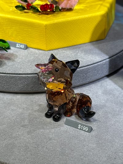 This image depicts a retail display in the city of Paradise, United States. The central subject is a faceted crystal figurine of a fox, predominantly amber-brown, with black ears, nose, feet, and a black-tipped tail. The fox is positioned on a grey, textured display surface. A grey price tag displaying "$199" in white text is situated in front of the fox. In the background, a curved grey display platform extends to the left, and a bright yellow, textured, multi-sided object is visible behind it. A partially obscured green, faceted object is visible on the upper grey display platform to the left, alongside a grey price tag displaying "$249". No individuals are present in the scene.