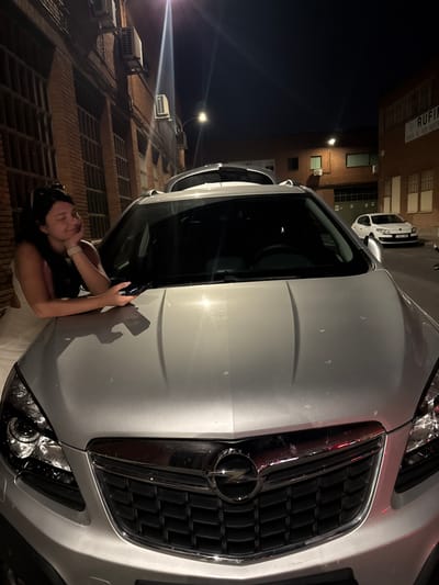 A young woman with dark hair is leaning on the hood of a silver Opel Mokka. She is wearing a white dress and has her eyes closed, appearing relaxed. She holds a smartphone in her right hand, resting it on the car's hood. The car is parked in front of a brick building at night. The building has large, barred windows and air conditioning units mounted on the exterior. Across the street, another building is visible with a sign that reads "RUFINO" and "VENTA AL POR MAYOR". A white Renault Megane is parked further down the street. Streetlights illuminate the scene.