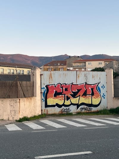 The image depicts a street scene in Piedrahíta, Spain, at what appears to be either dawn or dusk, indicated by the clear, pale blue sky and the warm light on distant mountains. In the immediate foreground, an asphalt road with a faded white pedestrian crosswalk extends horizontally. Above the crosswalk, a small strip of green vegetation grows at the base of a low, light-colored concrete wall, which transitions into a chain-link fence topped with barbed wire on the left side of the frame.
Dominating the midground is a large, corrugated metal gate, heavily rusted with a light blue-grey base peeking through extensive brown and orange discoloration. This gate is adorned with a prominent graffiti tag, featuring stylized, bubble-like letters filled with a gradient transitioning from red and orange at the top to yellow at the bottom, outlined in black with a thin white border. Small blue paint drips or dots flank the left and right edges of the graffiti. Legible text "2024." is visible below the graffiti on the left, and "SMARTIEVS" (or similar) on the right. The gate is supported by robust, light-colored concrete pillars on either side, with only the left pillar fully visible.
In the background, a cluster of buildings comprises various architectural styles typical of a Spanish town. To the far left, a multi-story structure with a light-colored facade and numerous windows is visible. Behind the gate, several houses feature terracotta-tiled roofs, light-colored walls, and multiple windows and balconies. Beyond the urban structures, a range of mountains, illuminated with a warm, reddish-brown hue, stretches across the horizon under the clear sky. No individuals or active interactions are present in the scene.