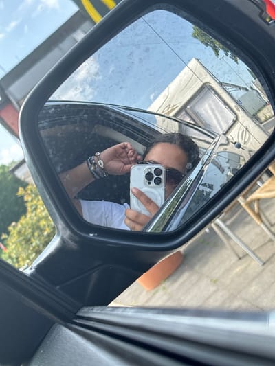 A person is taking a selfie in the side mirror of a vehicle. The reflection in the mirror shows the person holding a smartphone up to their face, wearing sunglasses and bracelets. The background reflection includes parts of another vehicle, a building, and foliage. It is not possible to confirm the location in Frechen, Germany, based on the visual information.