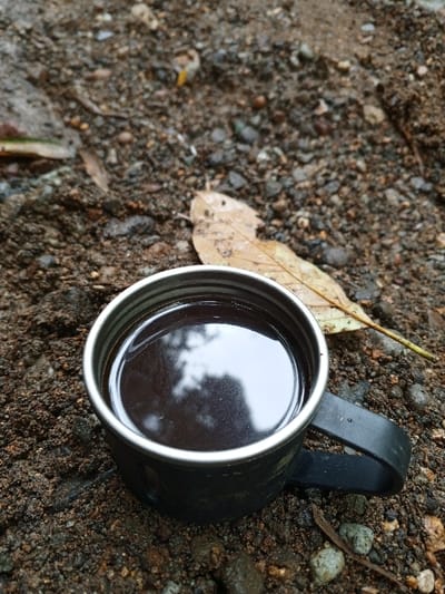 A dark-colored metal mug containing a dark liquid, presumed to be coffee, rests on damp, dark brown soil mixed with pebbles and small stones. The liquid surface reflects overhead light and indistinct shapes, possibly foliage. A single dry, light brown leaf is positioned behind and to the right of the mug on the ground. Additional small organic debris is scattered across the soil. No individuals are present. The scene is located outdoors in Silvino Lobos, Philippines.