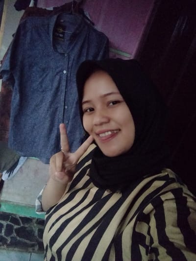A female individual is depicted in a close-up indoor portrait, positioned on the right side of the frame, within Lumajang, Indonesia. She wears a black hijab and a black and beige horizontally striped shirt. Her expression is a smile, with visible teeth and a partially extended tongue. Her right hand is raised to head level, forming a V-sign with the index and middle fingers; a ring is present on her right ring finger. In the background, to the left of the individual, a blue-grey short-sleeved button-up shirt with white buttons is hanging. A label bearing 'TOP BOY' is visible on the hanging shirt. The wall behind is painted with varying shades, including a light purple hue on the upper left. A patterned, dark-toned surface with a horizontal green stripe is visible on the lower left of the background. The lighting is moderate.