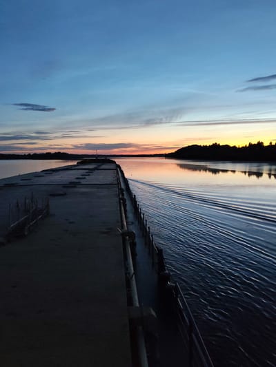 A concrete pier extends into a wide body of water under a twilight sky. The sky displays a gradient of colors, from deep blue at the top to orange and yellow near the horizon, with thin clouds streaking across. The water is calm, reflecting the sky's colors, and a noticeable wake trails behind the viewpoint, suggesting movement away from the pier. The pier itself is wide and flat, with a darker, textured surface. Along the edge of the pier, there are visible railings and some weathered wooden structures. The opposite bank of the water is lined with a dense, dark silhouette of trees. In the distance, a small navigational light is visible on the pier. The overall impression is one of peaceful solitude at dusk. The location is identified as Yakunina Gora, Russia.
