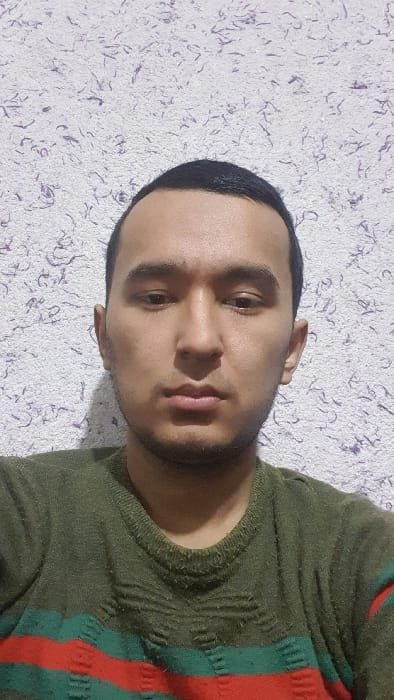 The image displays a male individual, appearing to be in his 20s or 30s, positioned centrally against a textured wall in what is identified as the city of Uchqo'rg'on, Uzbekistan. The subject has short, dark hair, a light mustache, and a short beard, with brown eyes looking directly into the camera, maintaining a neutral expression. He is wearing an olive green long-sleeved knit sweater featuring two prominent horizontal red stripes and two narrower teal-green stripes across the lower chest and abdomen; the sweater fabric exhibits visible pilling. The background wall is covered in a light-colored (possibly off-white or light grey) textured material, characterized by numerous small, irregular dark purple or dark grey fibers or flecks embedded within its surface. The scene is illuminated by an unseen indoor light source.