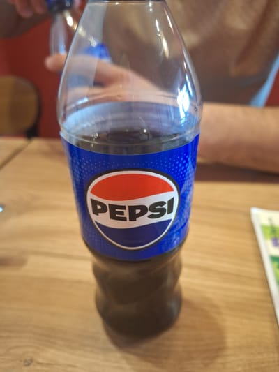 The image shows a close-up of a plastic bottle of Pepsi, with the blue and red Pepsi logo clearly visible on its label. The bottle is dark, filled with a dark liquid, and sits on a wooden table. In the background, a person's arm and shoulder are visible, suggesting they are holding or near the bottle. The lighting is bright, creating reflections on the plastic bottle. The context of Mińsk Mazowiecki, Poland is not discernible from the image content itself.