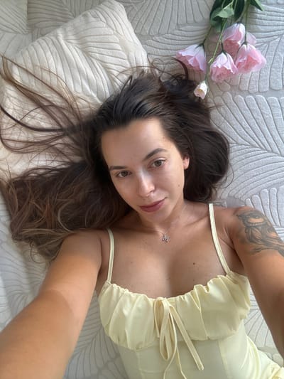 A woman with long, dark brown hair is lying on a textured, cream-colored surface, possibly a bed or a rug. Her hair is spread out around her head. She is wearing a light yellow, ruffled top with thin straps and a string tie at the bust. A silver necklace with a pendant shaped like the letter "M" is visible on her neck. On her left shoulder, she has a tattoo of an owl. To the upper right of her head, there are several light pink artificial flowers. Her body is positioned to the left of the frame, with her face slightly turned towards the camera. The environment appears to be indoors, with soft lighting. There is no specific information to determine the location in Torremolinos, Spain from the visual cues in the image.