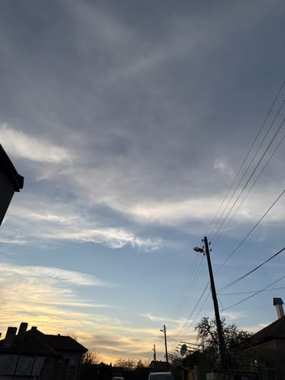 The image displays a residential street scene in Inovo, Bulgaria, observed at either dawn or dusk. The sky occupies the majority of the frame, transitioning from a dense layer of gray cloud cover in the upper half to a clearer, light blue sky with wispy white clouds and a distinct orange-yellow hue on the horizon to the left. Multiple utility poles, dark in silhouette, are present. A prominent pole in the mid-right features a single streetlight and supports numerous power lines that traverse diagonally across the frame. Additional poles and power lines extend into the background. Residential structures with dark-tiled roofs and chimneys are visible on both sides of the street, largely silhouetted against the horizon. Deciduous trees, appearing bare, are interspersed among the buildings and utility infrastructure. Several dark vehicle shapes are faintly discernible on the unlit street in the distance. No individuals or active interactions are present in the scene.