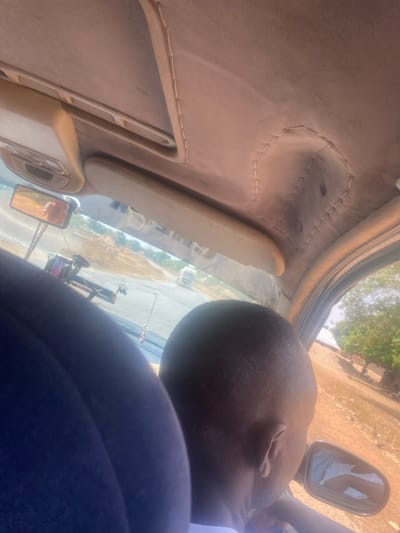 The scene is located within the city of Aboro, Nigeria. The image is captured from inside a moving vehicle, looking forward and slightly to the right. The vehicle's interior displays a beige headliner with visible stitching, an overhead console, and two beige sun visors. A rearview mirror reflects a partial image of a person's face. Suspended from the rearview mirror are a dark object, consistent with a phone mount, and a small, silver-colored hanging crucifix. The back of a person's head, with short-cropped dark hair, is prominently positioned on the right side of the frame, facing towards the vehicle's exterior right side mirror. A portion of this person's light-colored shirt is reflected in the side mirror. A dark blue front seat is partially visible on the left. Outside the vehicle, a paved road extends into the distance, bordered by reddish-brown dirt verges with sparse, dry vegetation. A light-colored vehicle, appearing to be a truck, is visible further down the road. In the background on the right, low-rise buildings with brown roofs are visible amidst trees, indicating a settlement. The scene is illuminated by strong daylight.