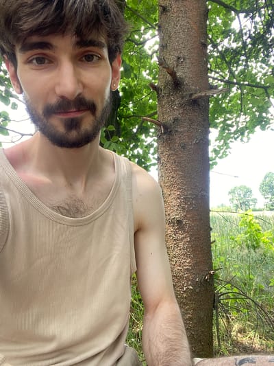 The image is a selfie taken outdoors, likely in Kaniów, Poland, given the context. A young man with brown hair and a beard is positioned in the foreground, looking directly at the camera. He is wearing a beige ribbed tank top. To his right, a thick tree trunk is visible, with some branches extending out. The background consists of lush green foliage and grass, with a hint of a field and distant trees. The lighting suggests it is daytime, with sunlight filtering through the leaves. The man's left arm is visible, and a tattoo can be seen on his left thigh. The overall scene depicts a casual outdoor setting.