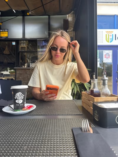 A young Caucasian woman with blonde hair and sunglasses is sitting at an outdoor table, holding an orange smartphone in her right hand. She is wearing a light yellow t-shirt. The setting appears to be a cafe or restaurant with a dark awning overhead and a textured grey placemat on the table.
In the foreground, a black and white "TO GO" coffee cup with a graphic of coffee beans is placed on a white saucer. There are also some red-wrapped items, possibly biscuits, on the saucer. A fork is partially visible peeking out from under a dark object to the right of the cup.
Behind the woman, a wooden crate holds two glass bottles, one filled with a dark liquid, likely syrup or coffee, and another with a clear liquid. In the background, there's a glimpse of a building with blue and white accents and some signage, suggesting an urban or town setting. The lighting indicates it is daytime.
The overall mood is casual and relaxed, with the woman appearing to be engaged with her phone. Visible text includes "TO GO" on the coffee cup, and some text on the building in the background, possibly in Spanish.