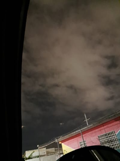 A nighttime exterior shot from inside a vehicle in Cumana, Venezuela, shows a multi-colored building with a red and yellow facade, featuring a white grid-like window. A utility pole with wires is visible in front of the building. The sky is filled with dense, dark clouds, illuminated by an unseen light source.