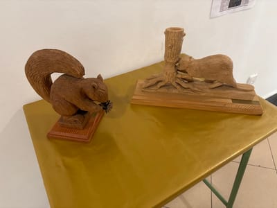 This image depicts two wooden animal carvings displayed on a table in an indoor setting within the city of El Espinar, Spain. The table is rectangular, covered with a shiny, crumpled gold-colored material, and has visible dark green metal legs.
On the left, a wooden sculpture of a squirrel stands on its hind legs, holding a detailed pinecone in its front paws. The squirrel has textured fur and a large, curved tail, and is mounted on a reddish-brown wooden base.
On the right, a wooden carving portrays a beaver gnawing on the base of a tree stump. The tree stump features exposed roots and a hollowed top, while the beaver is positioned low to the ground. This carving rests on a larger, light-colored wooden base with a small, rectangular brass-colored plaque affixed to its front.
The background consists of a plain white wall. In the upper right corner, a portion of a printed document, possibly an informational poster or newspaper clipping, is visible on the wall. The floor, partially visible in the bottom right, is composed of light-colored square tiles with dark grout.