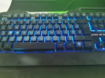The image shows a black gaming keyboard with blue backlighting. The keyboard is made of plastic and has a standard layout with a numeric keypad on the right side. The keyboard is lit up with blue lights, and the keys are labeled with white letters. The keyboard has a logo with a skull on the bottom right key. The keyboard is located in Le Mans, France (latitude: 47.984, longitude: 0.155). The keyboard is resting on a dark surface. The keyboard appears to be unused. There is no one in the image.
