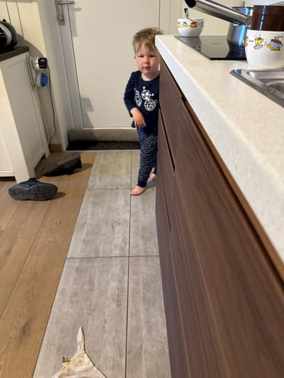 The image, rotated 90 degrees clockwise, depicts a domestic scene, likely a kitchen or an area adjacent to one. A young child with light brown hair, wearing a dark blue long-sleeved shirt with a white graphic and patterned pants, is visible in the mid-ground, looking directly at the camera with a slight smile or curious expression. The child appears to be standing upright on the floor.
In the upper portion of the corrected image, a white kitchen countertop with a built-in stainless steel sink and a modern faucet is prominent. Two white mugs, decorated with cartoon coffee/tea cup designs, sit on the countertop near the sink. Below the countertop are dark wooden cabinets or drawers, characterized by subtle horizontal handle grooves.
The floor transitions from light wooden planks in the foreground to grey tiled squares further back. Two dark-colored sneakers are placed on the wooden section of the floor. Partially visible on the tiled floor is a small, light-colored mat or rug, possibly animal-shaped. On a lower white cabinet or shelf against the wall, a black device (possibly a coffee maker or grinder), a power strip, a black adapter, and a small device with a blue light are arranged. A plain white wall or door is visible in the background. The general lighting suggests it is daytime. No discernible text is visible on any objects.