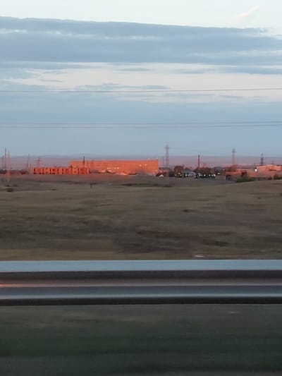 The image shows a landscape with a distant industrial complex under a cloudy sky. In the foreground, there is a grassy field with a horizontal railing in front. The industrial complex features several large buildings, some of which are illuminated with orange light, suggesting it might be dawn or dusk. Power lines and pylons are visible across the landscape. The overall scene is somewhat hazy, possibly due to atmospheric conditions or the quality of the photograph. There are no people or discernible actions in the image. The location is identified as Ayagöz, Kazakhstan.