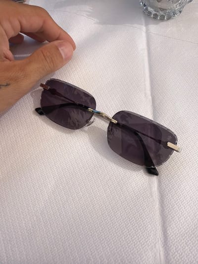 A pair of dark-lensed, rimless sunglasses with a silver frame rests on a textured white surface. The sunglasses are angled, with the left lens positioned slightly higher than the right. A thumb and part of a hand are visible in the lower left corner, holding the sunglasses. The hand appears to have a tattoo on the wrist. To the far left, a partially visible, ornate clear glass object, possibly a decorative bowl or dish, sits on the same surface. The background is a white, textured material, possibly a tablecloth or paper. The lighting suggests an indoor setting with overhead light.