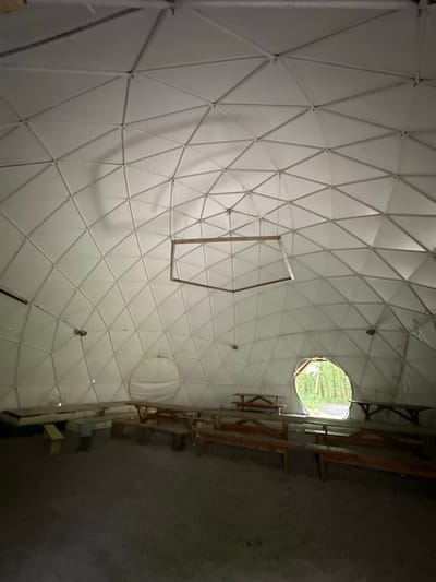 The image shows the interior of a geodesic dome. The dome's white, triangular panels form a complex geometric pattern on the ceiling. In the center of the dome, a wooden, hexagonal frame hangs from the ceiling by wires. Several wooden picnic tables and benches are arranged on the gravel floor. In the background, a large, oval opening in the dome reveals a view of a lush green forest. The lighting suggests it is daytime, but the interior is dimly lit, with light filtering in from the forest opening and possibly other unseen sources. The overall impression is of a rustic, communal space within a natural setting. This location is identified as the "Geodesic Dome at the Deerpark Forest Park" in Castlecomer, Ireland.
