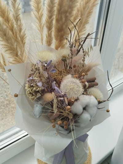 A dried flower bouquet is displayed on a windowsill in Bezlesnyy, Russia. The bouquet, composed of various dried plants such as pampas grass, cotton, dried lavender, and other seed pods, is wrapped in white paper and tied with a purple ribbon. The bouquet is placed in a woven basket, partially visible at the bottom of the frame. The background shows a window with a white frame and blinds, and through the glass, an outdoor environment is visible, suggesting a daytime setting. The lighting is natural, originating from the window.