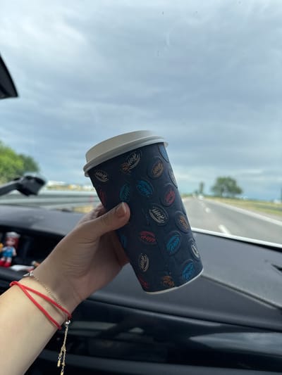 A person's hand holds a dark-colored disposable coffee cup inside a car, likely in Radinovo, Bulgaria. The cup is adorned with a repeating pattern of circular logos in various colors (blue, red, orange, beige) featuring the word "Happy" and the phrase "KEEP SMILING." The cup has a white rim. The hand, wearing two bracelets—a red string bracelet and a silver-toned chain bracelet with a small charm—is positioned in the lower right of the frame.
The setting appears to be the interior of a vehicle, with parts of the dashboard and windshield visible. Through the windshield, a bright, hazy sky is visible, indicating daytime. A blurred outdoor scene suggests a road or highway with distant trees and potentially a field. The overall impression is that of a journey or a stop along a route. The image is taken at eye level, looking out from the car.