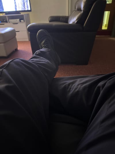 A first-person perspective photograph depicts an individual's lower body within an interior setting in Grays, United Kingdom. The individual is seated or reclined, wearing dark trousers and a dark lace-up boot on their right foot, which is elevated and angled towards the left. The left leg, also in dark trousers, is visible to the lower right.
The background reveals a room furnished with a dark brown or black leather recliner chair positioned centrally to the right. To the left, a white footstool or ottoman is partially visible. In the upper left corner, a flat-screen television displays a monochromatic image, possibly of a person. Beneath the television is a white multi-drawer cabinet, upon which a white electronic device, resembling a gaming console, is placed. The floor is covered with a reddish-brown carpet. Walls appear light-colored, and a doorway or opening in the far right background shows a sliver of yellow light. Artificial lighting illuminates parts of the room, casting shadows in the foreground.
