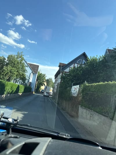 The media file captures a view from inside a vehicle, looking out at a residential street in Volpertshausen, Germany. The image is oriented vertically, suggesting it might have been taken while driving or is a rotated photograph.
The scene depicts a suburban street lined with houses and lush greenery. On the left side of the road, a light blue house with a dark brown roof is visible, surrounded by mature trees and manicured hedges. Further down the street, another house with a dark gray mansard roof and white trim is partially visible. A white sign with what appears to be an illustration is attached to a fence in front of this house.
On the right side of the street, a paved road stretches into the distance, with a light-colored sidewalk or path running alongside it. Several vehicles are parked or driving on the street. The weather appears to be sunny with a clear blue sky dotted with white, fluffy clouds. The bright sunlight suggests it is daytime, likely mid-morning or afternoon.
Reflected in the vehicle's windshield, a faint image of a person's hand can be seen, possibly holding the camera or interacting with the dashboard. The interior of the vehicle is also visible in the foreground, showing a dark dashboard and a device mounted on it. The overall atmosphere is peaceful and serene, capturing a typical day in a German village. No discernible text is clearly legible in the image, other than what might be on the partially visible sign.