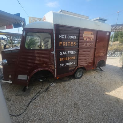 The image shows a maroon food truck parked on a gravel lot in La Palme, France, on a sunny day. The truck has white lettering on its side that reads "HOT DOGS," "FRITES," "GAUFRES," "BOISSONS," and "CHURROS." Below these words, it also says "LE SPOT." A small illustration of a truck is present near the text.
In the background, there are trees, sand dunes, and a wooden structure that appears to be part of an outdoor seating area or a pergola. There are also some people visible in the distance, but they are too far away to discern any details. Electrical cables are visible on the ground near the food truck, suggesting it is connected to a power source. The truck itself appears to be a vintage model, with a rounded front and a ribbed body. The overall impression is that of a casual outdoor dining establishment.