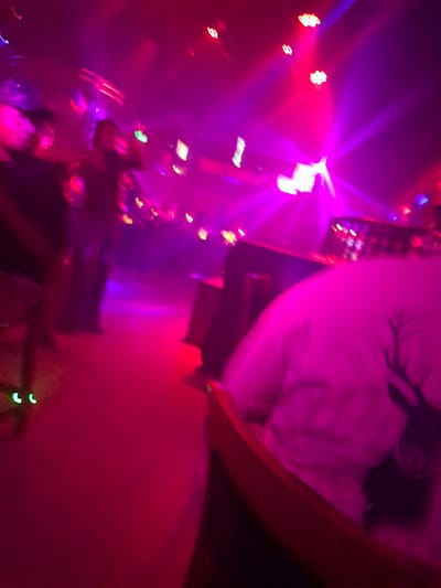 The media file depicts a dimly lit, vibrant nightclub scene in Nanning, China. The dominant colors are deep purples and vibrant pinks, cast by numerous lights that create a dynamic, atmospheric effect. In the foreground, a person is wearing a white t-shirt with a graphic of a deer's head. The t-shirt is partially obscured by the low-angle perspective and the pervasive lighting.
Several people are visible in the midground and background, though their features are indistinct due to the low light and motion blur, suggesting an active environment. Some individuals appear to be standing and possibly conversing, while others are less clearly defined. The setting includes what appears to be a bar area or elevated platform with lights and what might be bottles or glasses. The floor is a dark, reflective surface, likely a dance floor, illuminated by the ambient lights.
The activity taking place is consistent with a nightclub or similar entertainment venue, characterized by music, social interaction, and possibly dancing, although movement is difficult to discern clearly. The lighting is the most prominent feature, with beams and washes of color creating a lively and energetic mood. The time of day is clearly night. No specific weather conditions can be inferred. The location cues are limited to the context provided, which specifies Nanning, China. The overall emotion conveyed is one of excitement and a lively social atmosphere. There is no visible text in the media file.