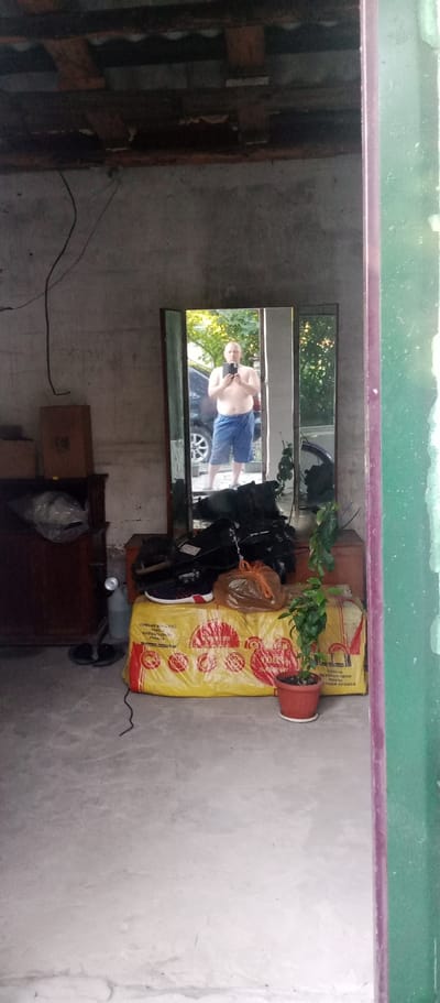 A full-body mirror is set up in what appears to be a garage or storage area, reflecting a man standing in front of it. He is shirtless and wearing blue shorts, holding a phone to take a selfie. The background visible in the mirror shows trees and a car, suggesting an outdoor setting beyond the garage entrance.

The garage itself is cluttered with various items. To the left, there is a dark wooden cabinet with items wrapped in plastic on top. A tall, clear plastic bottle with a silver cap stands next to it. In front of the mirror, a large yellow bag with red circular designs and some Cyrillic text is visible, with a pair of athletic shoes and a brown plastic bag on top. A small potted plant with green leaves sits on the floor to the right of the yellow bag.

The walls of the garage are made of concrete blocks, and the ceiling shows exposed wooden beams. The overall impression is of a somewhat disorganized, utilitarian space. The lighting suggests it is daytime, possibly late afternoon, given the warm light filtering through the trees in the mirror's reflection. The scene appears to be in Мариновка, Kazakhstan. The text on the yellow bag includes Cyrillic characters, but the specific words are not clearly legible.