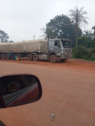 A large, grey truck is parked on a reddish-brown dirt road in Imilike, Nigeria. The truck is covered with a light-colored tarp. Three men are standing near the rear of the truck, and one of them appears to be pouring liquid from a yellow container. The truck has multiple wheels and appears to be a transport vehicle. In the foreground, a red side-view mirror of a car is visible, reflecting a person wearing a black t-shirt. The background consists of lush green trees and a cloudy sky.