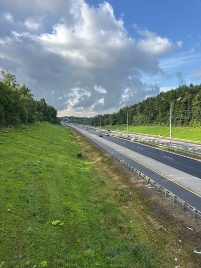 The image shows a highway with several cars driving on it, bordered by a steep, grassy embankment on the left and a dense forest on the right. The sky is partly cloudy with a mix of blue patches and heavy gray clouds. Streetlights line the highway, and a metal guardrail runs along the edge. The overall scene depicts a semi-urban or rural highway setting.