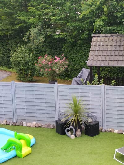 The image depicts a backyard in Troisdorf, Germany. In the foreground, an inflatable blue and green crocodile-shaped pool is visible on artificial turf. Behind the pool, a pale gray horizontal slat fence stretches across the image. To the right of the fence, two black storage boxes are placed on the turf, each topped with decorative gray hearts. A spiky potted plant sits on the left box. In the background, a dense green hedge forms a backdrop, with a trellised structure covered by a gray tarp partially visible on the right. A flowering bush with pink blossoms stands to the left of the trellised structure.