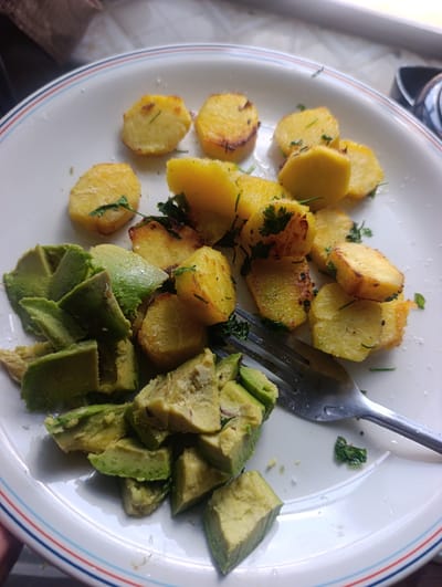 A close-up, overhead shot shows a white plate with a blue and red rim filled with roasted potato slices and cubed avocado. The potatoes are golden brown, some with slightly charred edges, and sprinkled with chopped green herbs. The avocado is bright green and appears fresh. A silver fork rests on the plate, partially submerged in the avocado. The background is blurred, suggesting an indoor setting. The scene is located in Dundori ward, Kenya.