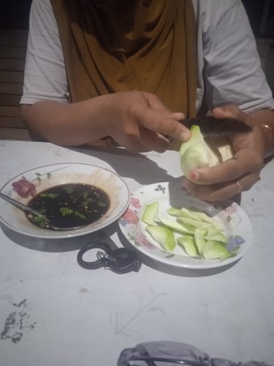 The image shows a person in a white t-shirt and a mustard-colored garment preparing a green fruit, likely a mango, on a white, patterned tablecloth. The person is using a knife to peel the fruit, with sliced pieces already placed on a small plate with floral decorations. To the left, another bowl holds a dark dipping sauce with what appear to be chopped green chilies. A spoon is in the sauce bowl. A set of keys with a black ring is on the table near the bowls. The setting appears to be indoors, possibly a home, with a wooden slatted surface visible in the background, suggesting it might be a porch or an outdoor covered area. The lighting is dim, indicating it could be evening or a poorly lit indoor space. The overall scene depicts a casual meal preparation, likely a snack, given the presence of the fruit and dipping sauce. The context provided indicates the location is Meunasahblang, Indonesia. No visible text is present in the image.