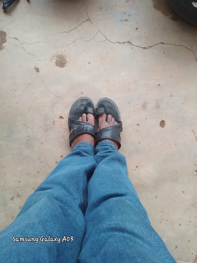 A low-angle, medium shot captures a person's legs from the knees down, wearing blue denim jeans and black sandals with a textured, crocodile-like pattern. The sandals have a thong design, with the big toe passing through a strap. The person is standing on a cracked concrete surface, possibly a road or courtyard, with scattered dark spots and lighter patches of discoloration. A faint watermark in the bottom left corner reads "Samsung Galaxy A03." The environment appears to be outdoors, with an uneven and worn-out ground. The overall impression is one of an everyday, unposed moment captured in a somewhat neglected urban or semi-urban setting.