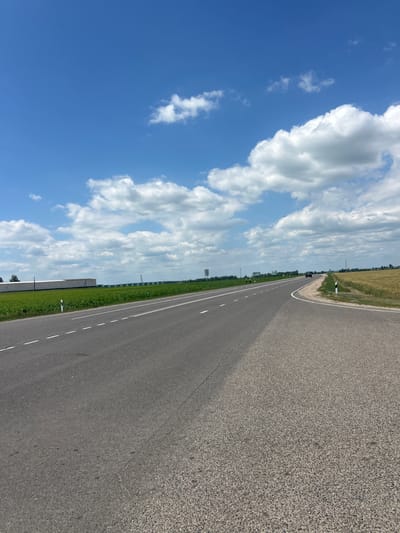This media file captures a view of an asphalt road curving to the right, bordered by green fields and a clear blue sky with white, fluffy clouds. The scene is outdoors, suggesting a rural or roadside setting, possibly in Belarus given the provided location context. The perspective is from the side of the road, looking forward and slightly upwards.
The road itself is gray asphalt with white dashed lines indicating lanes. A single dark vehicle is visible in the distance on the road. Along the side of the road, there are patches of green grass and vegetation, with some taller greenery suggesting agricultural fields or a treeline further back. There are also a few white roadside markers.
The sky is a vibrant blue, filled with cumulus clouds that are bright white and appear to be lit by the sun. This indicates it's daytime, likely midday or afternoon, given the brightness and the shadows cast on the road. The weather appears to be clear and pleasant.
No people are clearly visible in the scene. The overall mood is serene and expansive, with the open road and sky evoking a sense of journey or travel. The visual elements are simple and natural, dominated by the road, sky, and greenery. There is no visible text within the image.