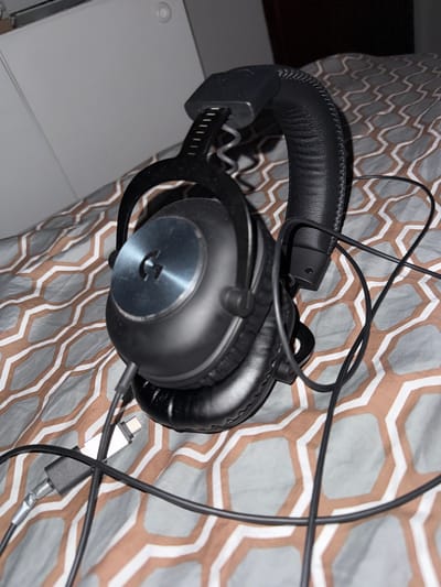This media file shows a close-up of a pair of black Logitech gaming headphones resting on a patterned comforter. The headphones are the primary object in focus, with their leather headband and earcups clearly visible. Wires from the headphones are also visible, some neatly coiled and others trailing across the comforter. One of the wires appears to be connected to an adapter.
The background is a comforter with a repeating hexagonal pattern in shades of gray and brown. The lighting suggests it is indoors, possibly in a bedroom, during the daytime. There are no people visible in the image, and the scene appears to be static, with the headphones simply placed on the bed.
A notable detail is the Logitech logo, a stylized "G" in a silver circle, on the side of one of the earcups. The overall impression is one of a personal electronic device within a domestic setting. The image is taken from a slightly elevated angle, looking down onto the headphones and comforter. There is no visible text other than the brand logo on the headphones.