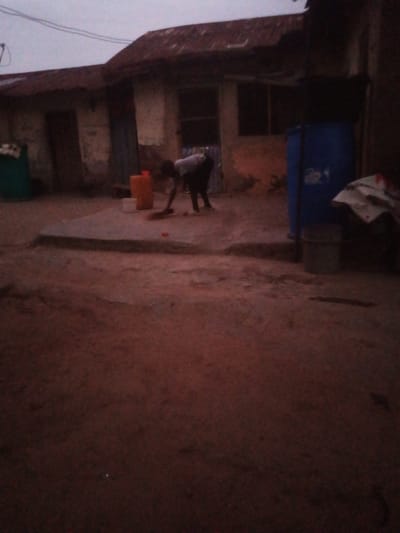 The scene shows an individual sweeping an outdoor courtyard in Kupai, Nigeria. The courtyard is partially paved with a concrete slab, and the surrounding ground appears to be dirt. Various containers are visible, including orange, white, and blue barrels or buckets. To the left, a green bin stands near the entrance to a weathered building with a corrugated metal roof. The individual is bent over, sweeping the concrete slab in front of the building. The light suggests it is either dusk or dawn.