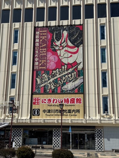 The image shows a multi-story building in Nakatsugawa, Japan, featuring a large advertisement for "Jikabuki", referring to local traditional kabuki theater. The advertisement depicts a kabuki mask and Japanese script. Adjacent to the kabuki ad is signage for the "Nakatugawa Souvenir Tourist Information Center". The building's architecture includes a facade with rows of windows and a decorative lattice design around the entrance. A street sign is visible near the entrance. The building appears to be located on a street with trees.