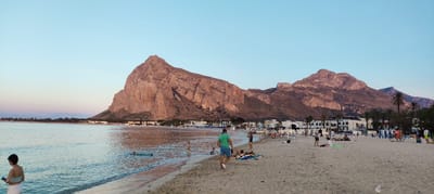 This is a panoramic photograph of a beach scene at what appears to be dusk or early evening. The sky is a soft gradient of blue and pale pink. The foreground features a sandy beach with several people engaged in various activities. Some individuals are walking along the shore, while others are relaxing on towels, sunbathing, or swimming in the calm, shallow water. A prominent natural feature in the background is a large, rugged mountain that dominates the left side of the frame. Additional, smaller mountains are visible further to the right. Along the coastline, there is a town with white buildings and some palm trees. The overall atmosphere is serene and relaxed, suggesting a pleasant summer evening by the sea. There is some visible text on a building in the background which appears to be "HOTEL [illegible]" or similar.