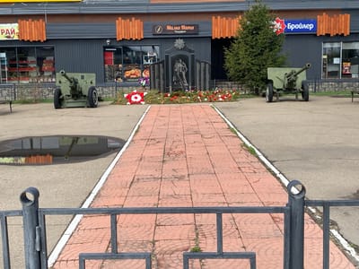 The image captures a memorial in Oyok, Russia. The memorial is flanked by two green cannons, positioned symmetrically on either side of a central pathway. The pathway is paved with red bricks, bordered by a white line. In the background, a dark gray building with signs in Russian: "Рай", "Мама Ната Пиццерия," and "Бройлер," which seem to indicate shops or restaurants. The memorial itself is decorated with wreaths and flowers. The scene is viewed through a dark gray metal fence. A puddle reflects a building in the left foreground. The weather appears to be overcast.