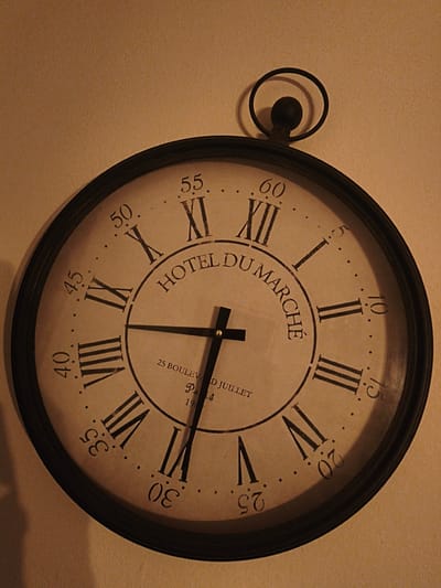 The image shows a large, round clock with a black frame hanging on a beige wall. The clock face is light brown with black Roman numerals indicating the hours. The minute markers are represented by small dots around the perimeter, with numerals indicating the minutes at intervals of five. The clock hands are black; the minute hand is pointing towards the 31-minute mark, and the hour hand is pointing towards the 7-hour mark.

In the center of the clock face, it reads "HOTEL DU MARCHÉ" in a decorative font, with "25 BOULEVAD JUILLET Paris 1904" printed underneath. The clock has a decorative ring and knob at the top, giving it the appearance of a pocket watch. The photo is taken indoors, and the lighting casts a warm tone on the scene.