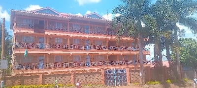 The image shows a three-story building with a peach-colored exterior and several balconies with iron railings. There are several windows on each floor and a large, brown wooden door at ground level. The building is surrounded by lush greenery, including palm trees, bushes, and shrubs. There is a small, black gate at the front of the building, and a dirt driveway leading up to it. The ground is a mixture of dirt and grass. The building appears to be located in a residential area, as there are other buildings in the background. The location of the building is likely on a street in Kanjuu, Kenya, with the coordinates -0.541 latitude and 37.441 longitude. There is a person walking towards the building in the bottom right corner of the frame. They are obscured by the tree in front of the building. The building is surrounded by a brick wall, and there is a small, brown door on the left side of the building. The building is located in a sunny area, and there is a blue sky with a few clouds in the background.