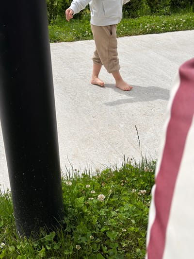 A child in tan pants and a white top stands barefoot on a concrete surface next to grass. The lower portion of a black cylindrical object is visible. A red and white striped fabric is in the background. The location is speculated to be Chkalov, Armenia.