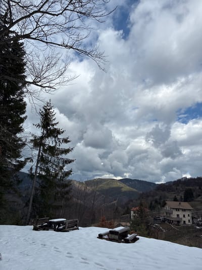 The image shows a mountainous landscape in winter. Dominating the upper portion are thick, grey clouds, punctuated with patches of blue sky. A hillside covered with trees, some evergreen and others bare, slopes upwards in the midground. A couple of buildings are perched on the hillside. In the foreground, a snow-covered surface contains the corner of a wooden table and chairs. There is no verifiable evidence that the location is Manastir, Bulgaria.