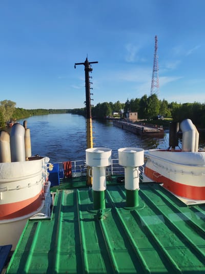 This image presents a sunny, clear day on a wide waterway, likely a river or canal, viewed from the deck of a moving vessel. The low angle of the sun, casting long shadows across the deck, suggests either morning or late afternoon. The sky is a vibrant blue with only a few wispy clouds.
In the immediate foreground, the vessel's green deck is prominent, adorned with white and red sections of the hull. Several large, silver-colored exhaust pipes are visible on the left, while a tall, dark mast-like structure rises centrally, equipped with various fittings. On a white cylindrical object on the deck, the Cyrillic word "АККУМУЛЯТОР" (ACCUMULATOR) is clearly legible, and "АвиаХимПром" (AviaKhimProm) can be faintly seen on another.
The vessel is moving forward, evidenced by the gentle ripples on the water's surface. Both banks of the waterway are lined with dense, lush green trees. In the middle distance on the right, a small building with a brown roof and a long, flat barge or pontoon docked alongside are visible, hinting at a navigational facility or mooring point. Further in the distance, a tall red and white lattice transmission tower stands out above the treeline. The scene depicts a peaceful moment of inland navigation, consistent with the provided location context of Annensky Most, Russia.