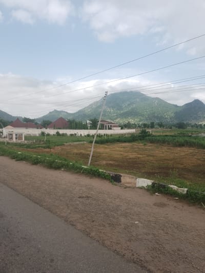 This media file depicts a landscape scene in Wuro Sani, Nigeria. The image showcases a vast expanse of land with rolling green hills in the background, indicative of a rural or semi-rural setting. Several buildings with distinctive red, conical roofs are visible on the left side of the frame, suggesting a residential or communal area. These structures are enclosed by a white wall, further defining the property.
In the foreground, a dirt road with a paved section on the left runs diagonally across the frame. The road appears to be a pathway or a rural track. Beside the road, there is a strip of green vegetation, transitioning into a dry, grassy field further into the landscape. Power lines are strung across the sky, supported by a tall pole located in the middle of the scene. The sky is partly cloudy, with a mix of blue sky and white clouds, suggesting it is daytime, possibly late morning or afternoon. The overall impression is one of a tranquil, somewhat undeveloped area, characteristic of many Nigerian landscapes. No people or specific activities are apparent in the image.
